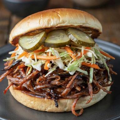 BBQ jackfruit sandwich on a toasted bun: sticky dark shredded jackfruit with charred edges under a vinegar slaw of green cabbage and carrot, topped with dill pickles