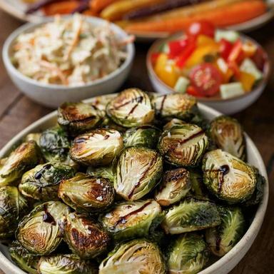 Four vegetable side dishes on a dark wooden table: a bowl of roasted Brussels sprouts with balsamic glaze in front, with whole roasted rainbow carrots, creamy coleslaw and a chopped pepper, cucumber and tomato salad behind.
