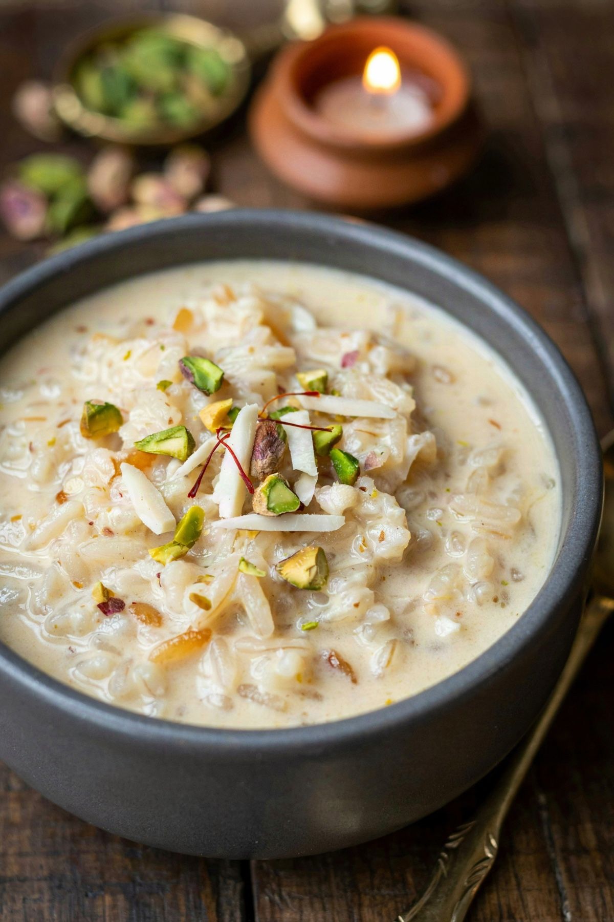 Kheer in a charcoal bowl, creamy pale ivory rice and milk pudding topped with slivered almonds, chopped pistachios and saffron threads, a lit clay lamp behind