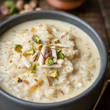 Kheer in a charcoal bowl, creamy pale ivory rice and milk pudding topped with slivered almonds, chopped pistachios and saffron threads, a lit clay lamp behind