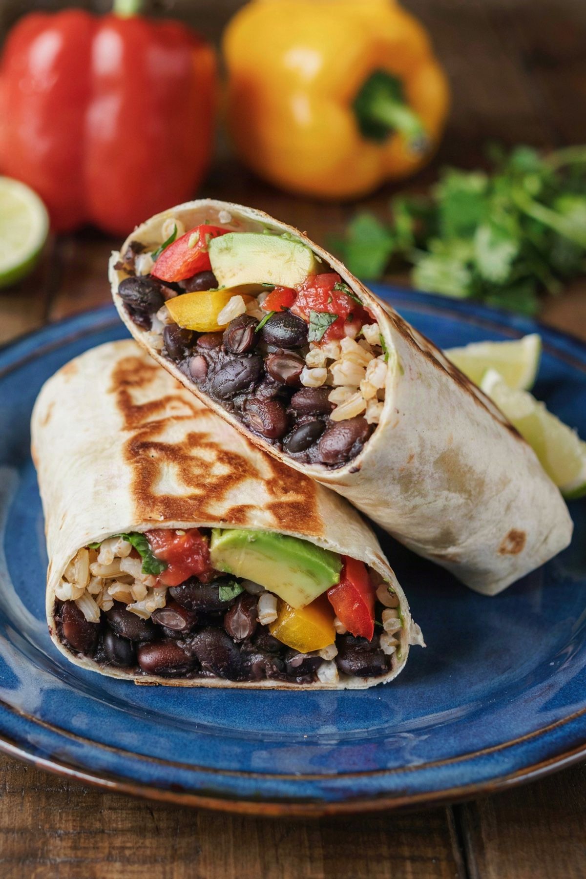 Vegan burrito cut in half on a cobalt-blue plate, a seared flour tortilla packed with black beans, brown rice, red and yellow pepper, avocado and salsa, with lime wedges
