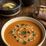 A wide bowl of butternut squash soup swirled with cream and scattered with pumpkin seeds and parsley, with bowls of black bean soup and tomato soup and torn crusty bread on the dark wooden table behind it.
