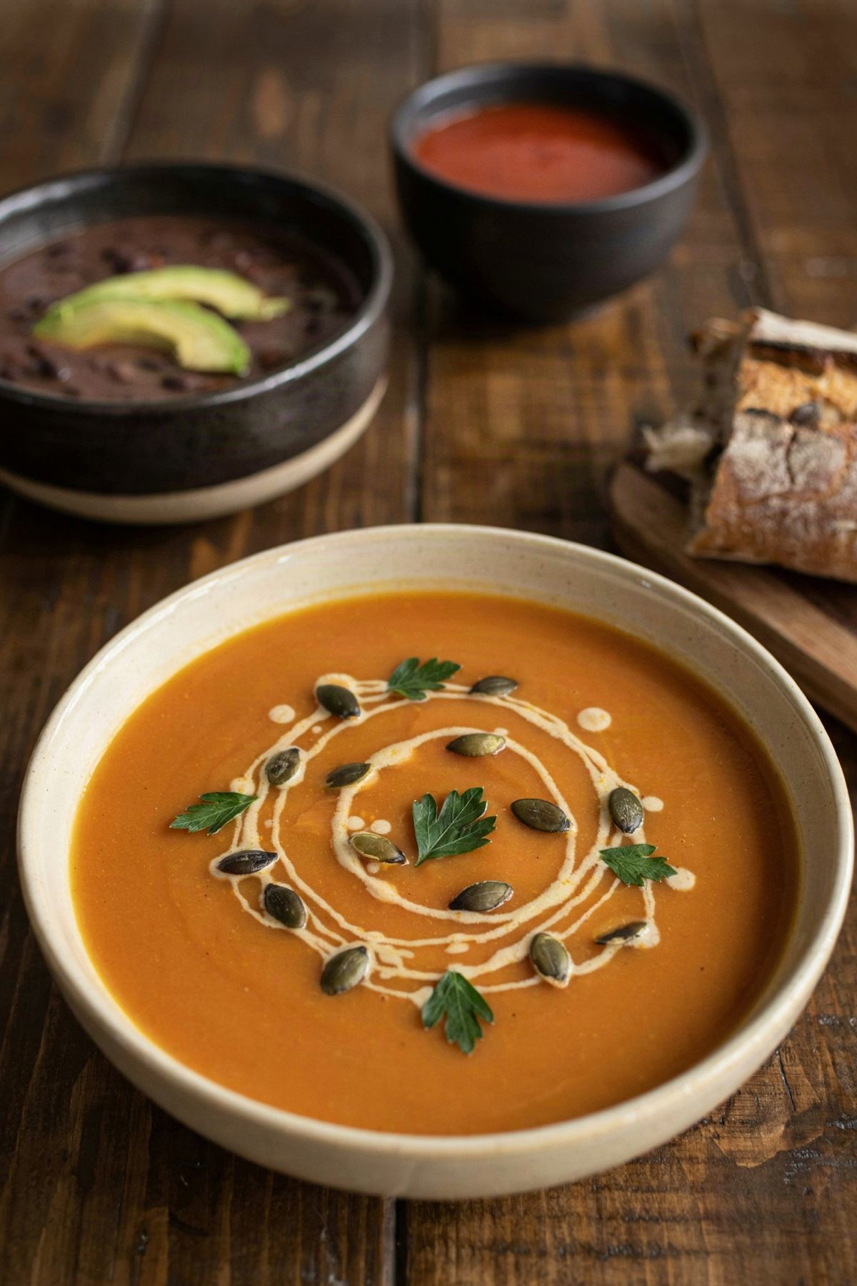 A wide bowl of butternut squash soup swirled with cream and scattered with pumpkin seeds and parsley, with bowls of black bean soup and tomato soup and torn crusty bread on the dark wooden table behind it.