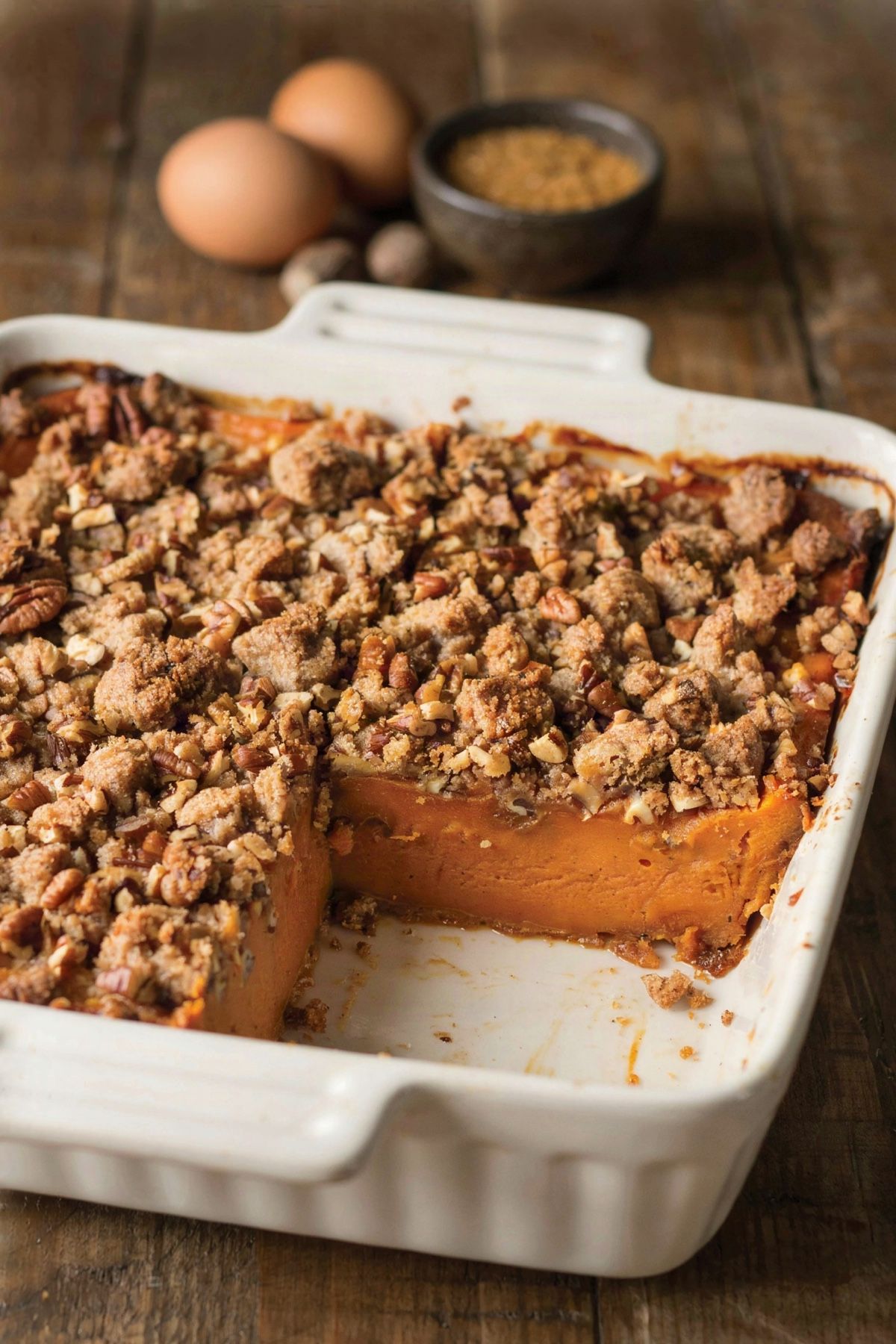 Sweet potato casserole in a white baking dish, a golden pecan streusel crust with one square lifted out to show the dense set orange filling underneath.