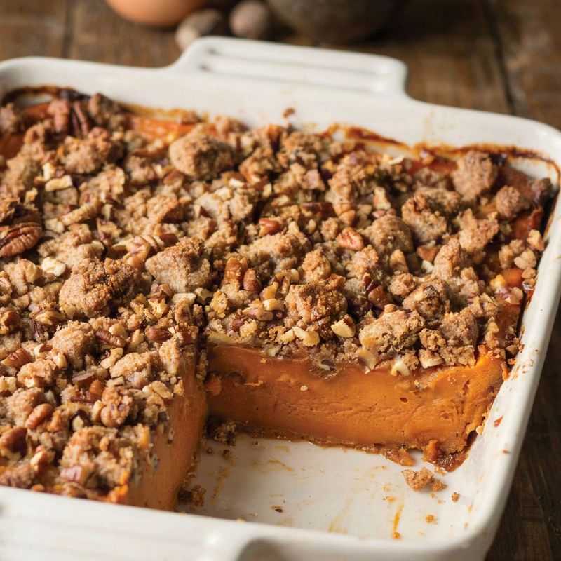 Sweet potato casserole in a white baking dish, a golden pecan streusel crust with one square lifted out to show the dense set orange filling underneath.