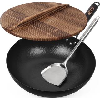Black hammered carbon steel flat-bottom wok with a steel handle, shown with a wooden lid and a stainless steel spatula