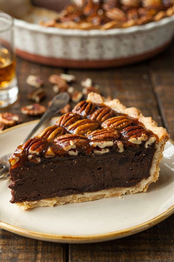 A slice of brownie pecan pie on a cream plate, its cut face showing a pale gold pastry base, a thick dark fudgy brownie layer and a glossy amber pecan layer under whole pecan halves.