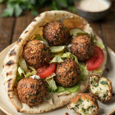 Falafel sandwich in a soft pita with tahini, craggy golden falafel, shredded lettuce, tomato and pickles, one falafel torn open beside it to show the green herb-flecked inside.