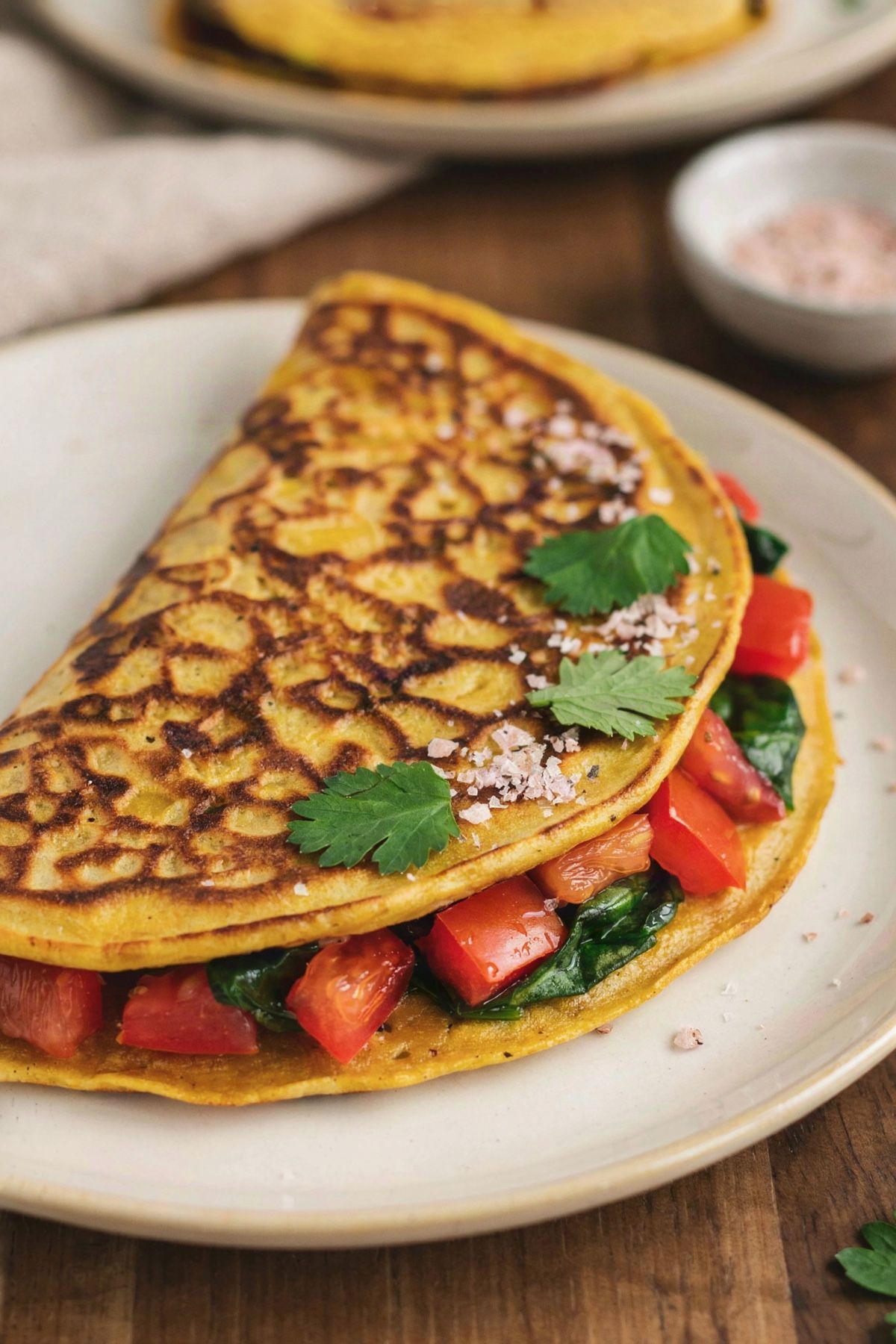 A folded vegan chickpea omelette on a pale ceramic plate, the golden turmeric-yellow pancake opened along one edge to show diced red bell pepper, tomato and wilted spinach, finished with a dusting of pink-gray kala namak and fresh cilantro.