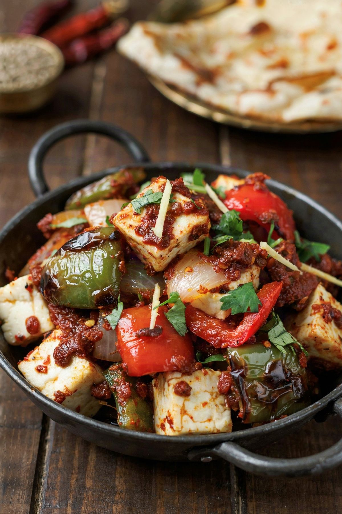 Kadai paneer in a black iron kadai, white paneer cubes and charred green and red pepper squares coated in a thick brick-red masala, topped with cilantro and ginger strips, naan behind
