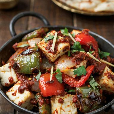 Kadai paneer in a black iron kadai, white paneer cubes and charred green and red pepper squares coated in a thick brick-red masala, topped with cilantro and ginger strips, naan behind