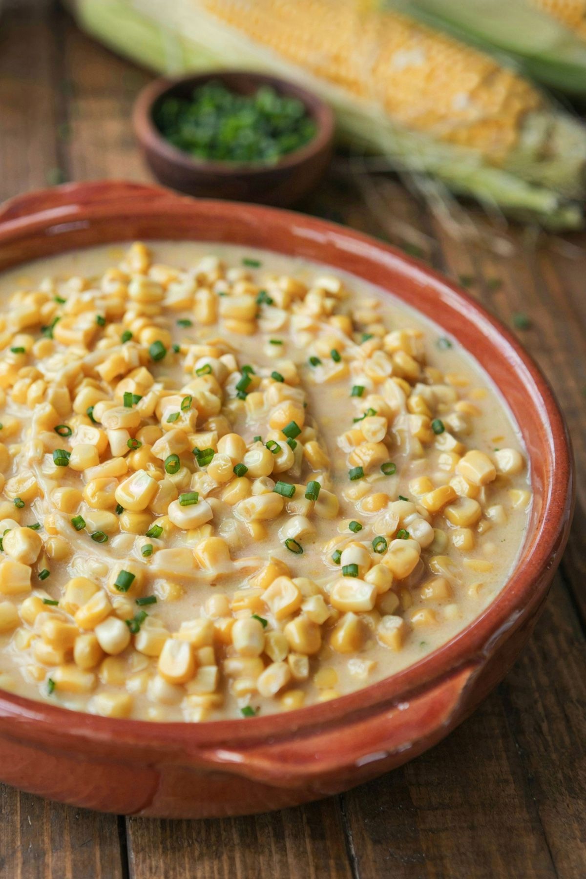 Vegan creamed corn in a rust-glazed stoneware bowl, whole sweet corn kernels suspended in a loose pale golden sauce and scattered with chopped chives, with husked ears of corn on the table behind.