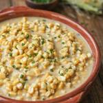 Vegan creamed corn in a rust-glazed stoneware bowl, whole sweet corn kernels suspended in a loose pale golden sauce and scattered with chopped chives, with husked ears of corn on the table behind.