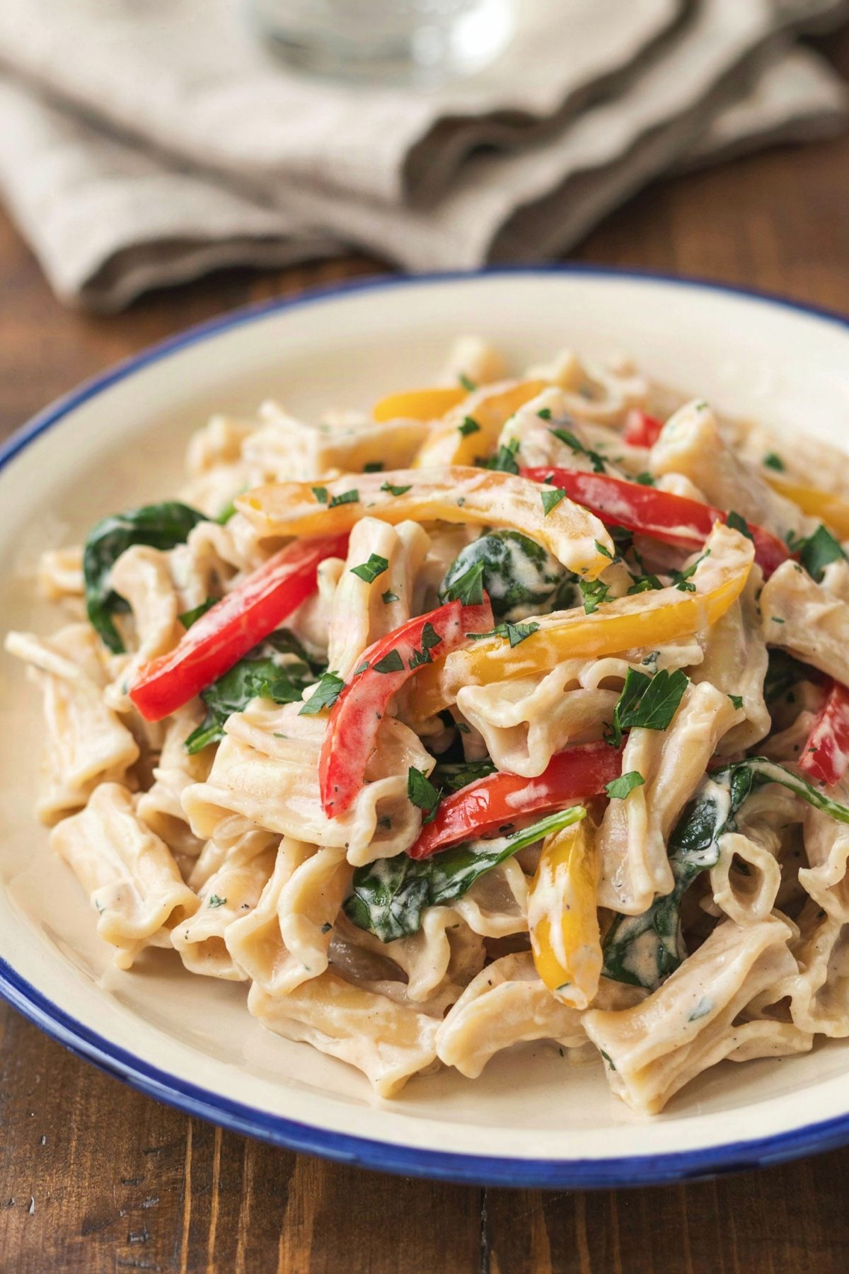 Ruffled campanelle thickly coated in a pale ivory tofu sauce, with red and yellow bell pepper strips, wilted spinach and chopped parsley, on a cream plate with a blue rim
