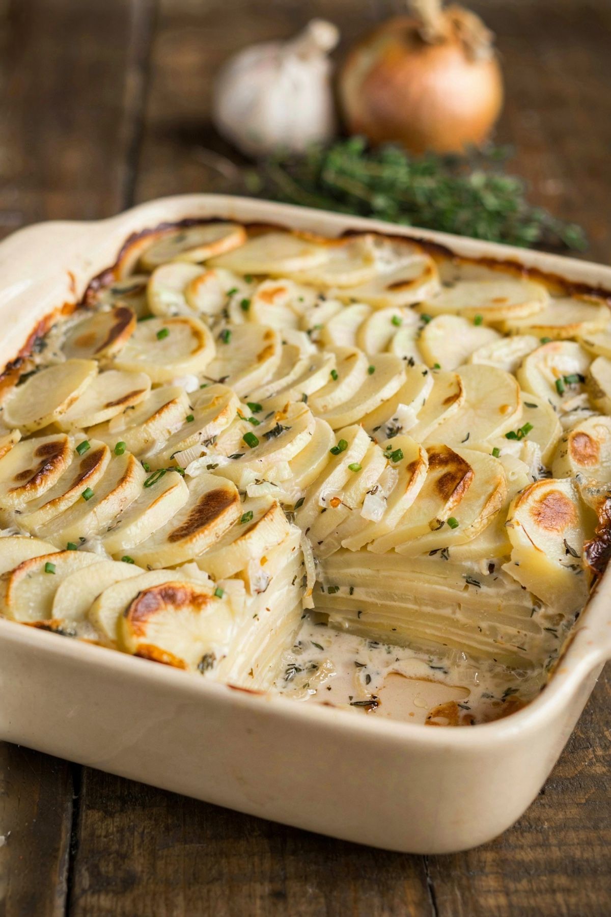 Vegan scalloped potatoes in a cream stoneware baking dish, wafer-thin rounds of potato in overlapping rows under a thick ivory sauce flecked with thyme, browned in patches on top and scattered with chives, with one corner portion lifted out to show a dozen thin layers inside.