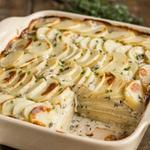 Vegan scalloped potatoes in a cream stoneware baking dish, wafer-thin rounds of potato in overlapping rows under a thick ivory sauce flecked with thyme, browned in patches on top and scattered with chives, with one corner portion lifted out to show a dozen thin layers inside.