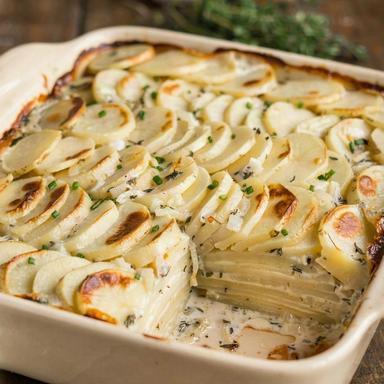 Vegan scalloped potatoes in a cream stoneware baking dish, wafer-thin rounds of potato in overlapping rows under a thick ivory sauce flecked with thyme, browned in patches on top and scattered with chives, with one corner portion lifted out to show a dozen thin layers inside.