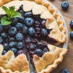 A golden brown vegan blueberry pie with a slice cut out, showcasing the juicy filling, on a rustic wooden table.