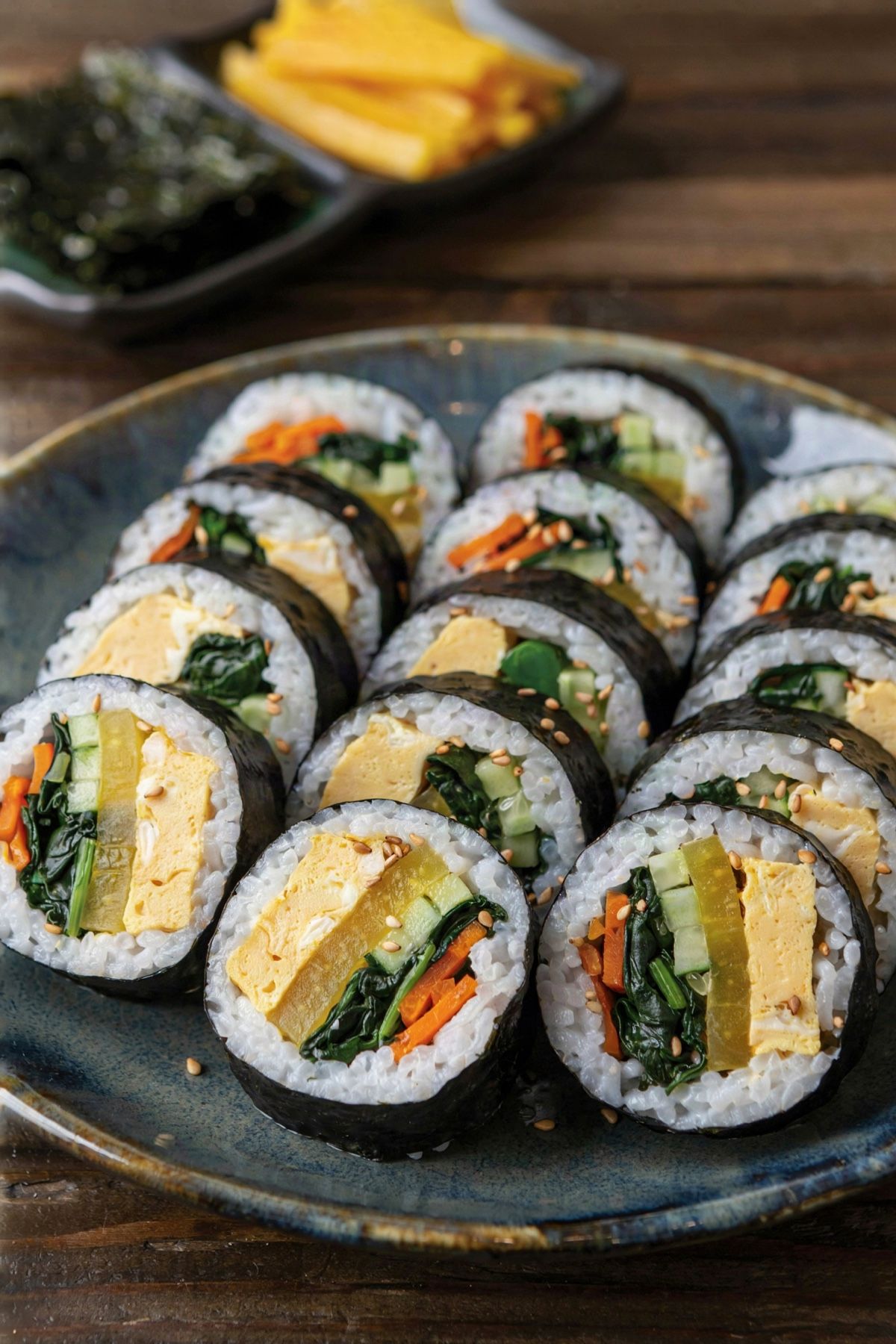 Vegetarian kimbap slices arranged cut side up on a dark ceramic platter, each showing egg, yellow pickled radish, spinach, carrot and cucumber inside a ring of white rice and black seaweed, scattered with sesame seeds.