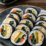Vegetarian kimbap slices arranged cut side up on a dark ceramic platter, each showing egg, yellow pickled radish, spinach, carrot and cucumber inside a ring of white rice and black seaweed, scattered with sesame seeds.
