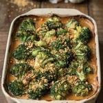 Vegan broccoli casserole in a cream stoneware baking dish, roasted broccoli florets with browned tips coated in a thick orange-gold sauce under a scattered layer of golden buttery cracker crumbs.