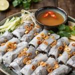 A plate of vegetarian banh cuon, thin translucent steamed rice rolls with a dark mushroom and wood ear filling showing through, scattered with golden fried shallots, with cucumber, bean sprouts and herbs and a small bowl of amber dipping sauce with red chili slices.