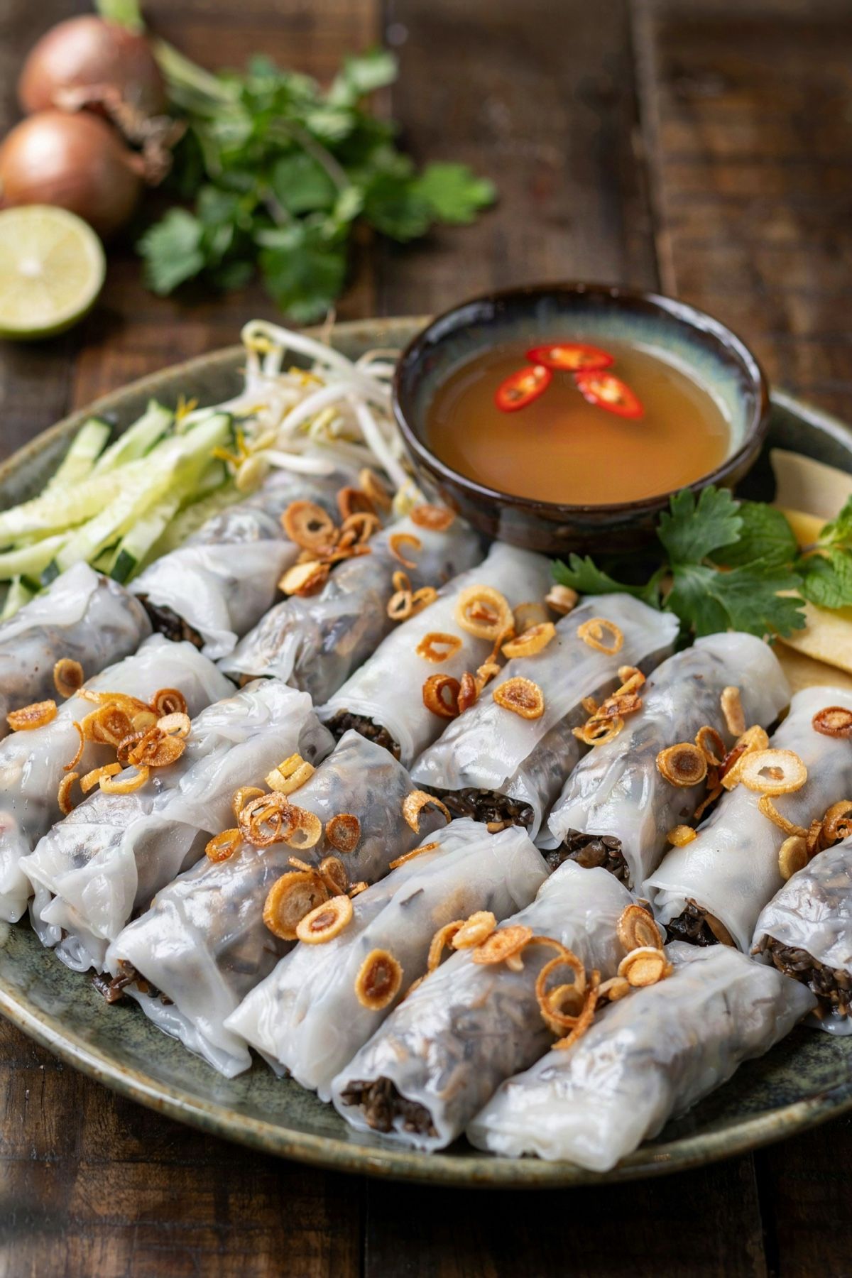 A plate of vegetarian banh cuon, thin translucent steamed rice rolls with a dark mushroom and wood ear filling showing through, scattered with golden fried shallots, with cucumber, bean sprouts and herbs and a small bowl of amber dipping sauce with red chili slices.