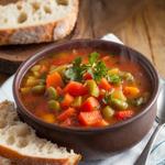 A bowl of hearty vegetable soup with colorful vegetables and parsley, accompanied by bread on a wooden table.