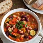 A hearty bowl of vegan minestrone soup with vegetables, beans, and pasta, garnished with parsley, on a rustic table with bread.