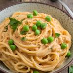 Vegan spaghetti carbonara with cashew sauce, peas, and parsley in a rustic bowl on a wooden table.