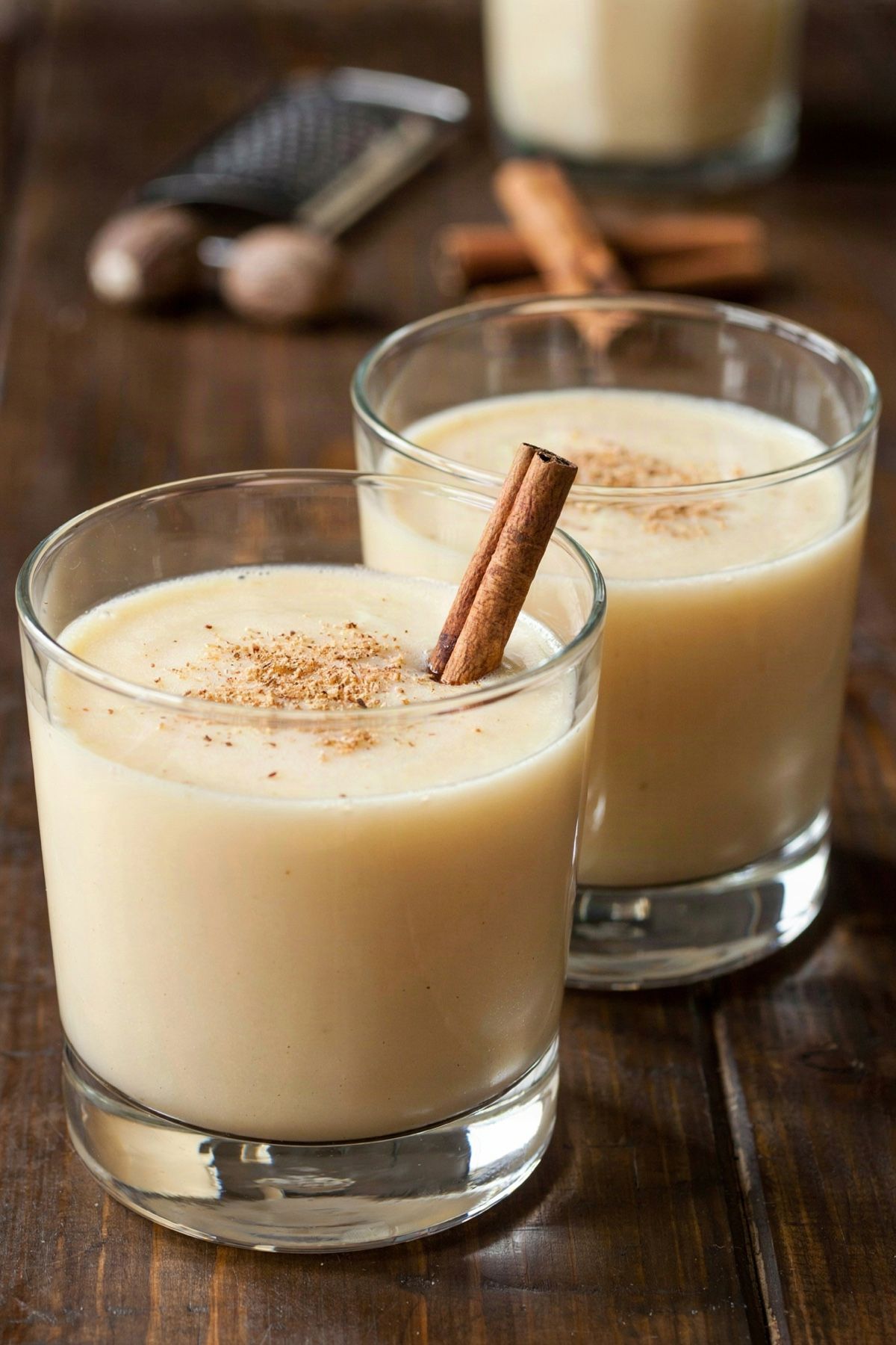 Two glasses of chilled vegan eggnog on a dark wooden table, a pale creamy cashew and coconut drink dusted with freshly grated nutmeg, one with a cinnamon stick, whole nutmegs and a grater behind