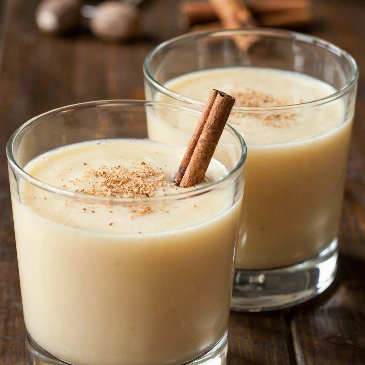 Two glasses of chilled vegan eggnog on a dark wooden table, a pale creamy cashew and coconut drink dusted with freshly grated nutmeg, one with a cinnamon stick, whole nutmegs and a grater behind