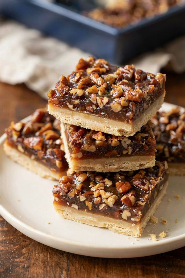 Pecan pie bars stacked on a cream plate, a dark amber filling packed with chopped pecans over a pale golden shortbread base