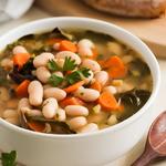 A bowl of vegan white bean soup with carrots and greens, garnished with parsley, on a rustic kitchen table with bread.