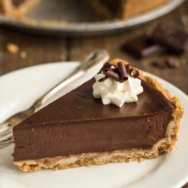 A slice of silken chocolate pie on a white plate, a dense dark chocolate layer on a graham cracker crumb base, topped with whipped cream and dark chocolate curls