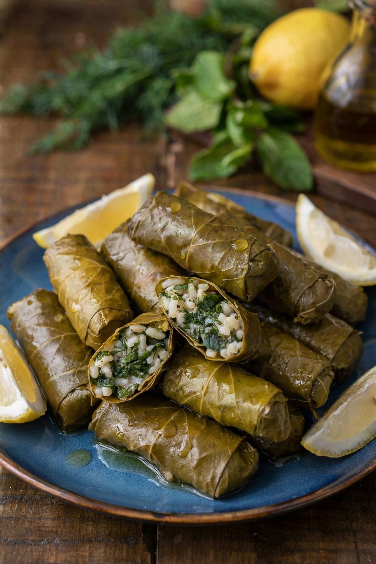 Greek dolmades piled on a blue plate, glossy rolled grape leaves with one cut open to show the green herb and rice filling, served with lemon wedges and a drizzle of olive oil.