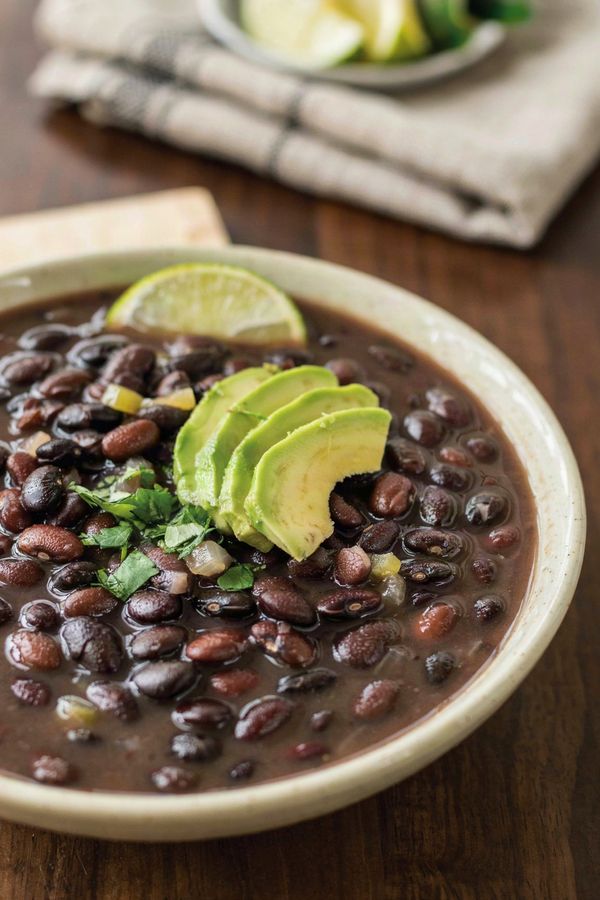 A cream ceramic bowl of dark black bean soup with whole black beans, topped with fanned avocado slices, chopped cilantro and a lime wedge, on a dark wooden table.