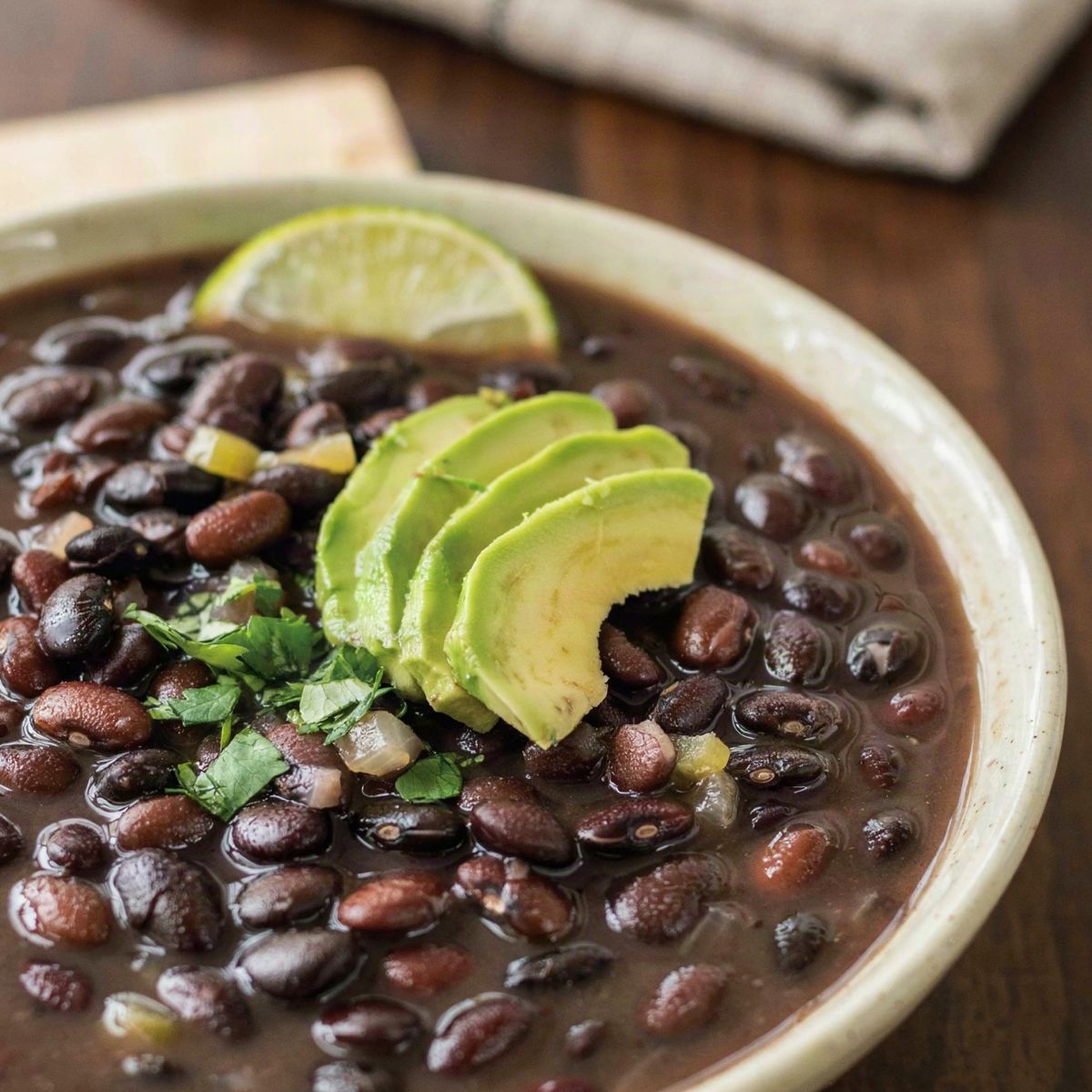 A cream ceramic bowl of dark black bean soup with whole black beans, topped with fanned avocado slices, chopped cilantro and a lime wedge, on a dark wooden table.
