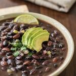 A cream ceramic bowl of dark black bean soup with whole black beans, topped with fanned avocado slices, chopped cilantro and a lime wedge, on a dark wooden table.