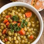 A bowl of meatless split pea soup with green peas, carrots, and celery, garnished with parsley, served with bread on a wooden table.
