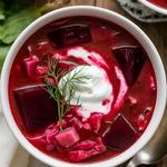 A bowl of vegetarian borscht soup with beets, potatoes, and cabbage, garnished with sour cream and dill on a rustic table.