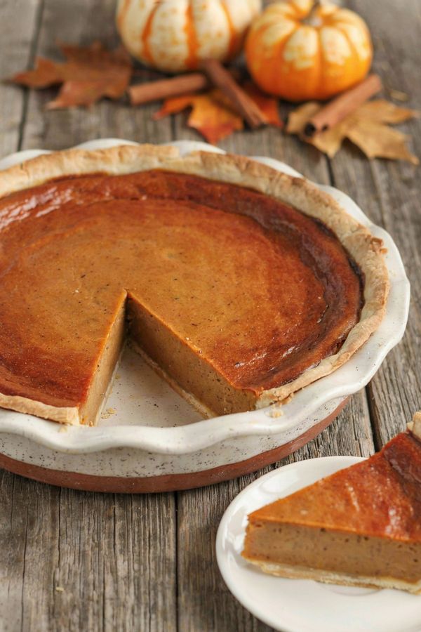Whole vegan pumpkin pie in a fluted ceramic dish with one wedge cut out and set on a plate, the deep orange filling firm and sliced clean, on a weathered wooden table with small pumpkins and autumn leaves.