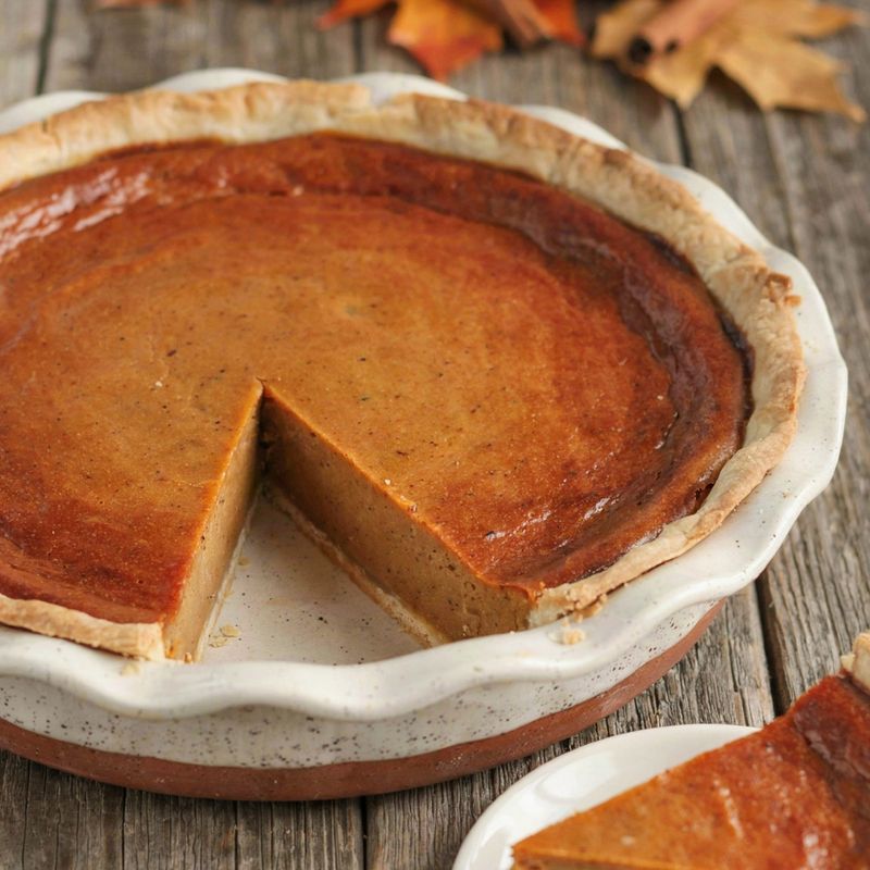 Whole vegan pumpkin pie in a fluted ceramic dish with one wedge cut out and set on a plate, the deep orange filling firm and sliced clean, on a weathered wooden table with small pumpkins and autumn leaves.