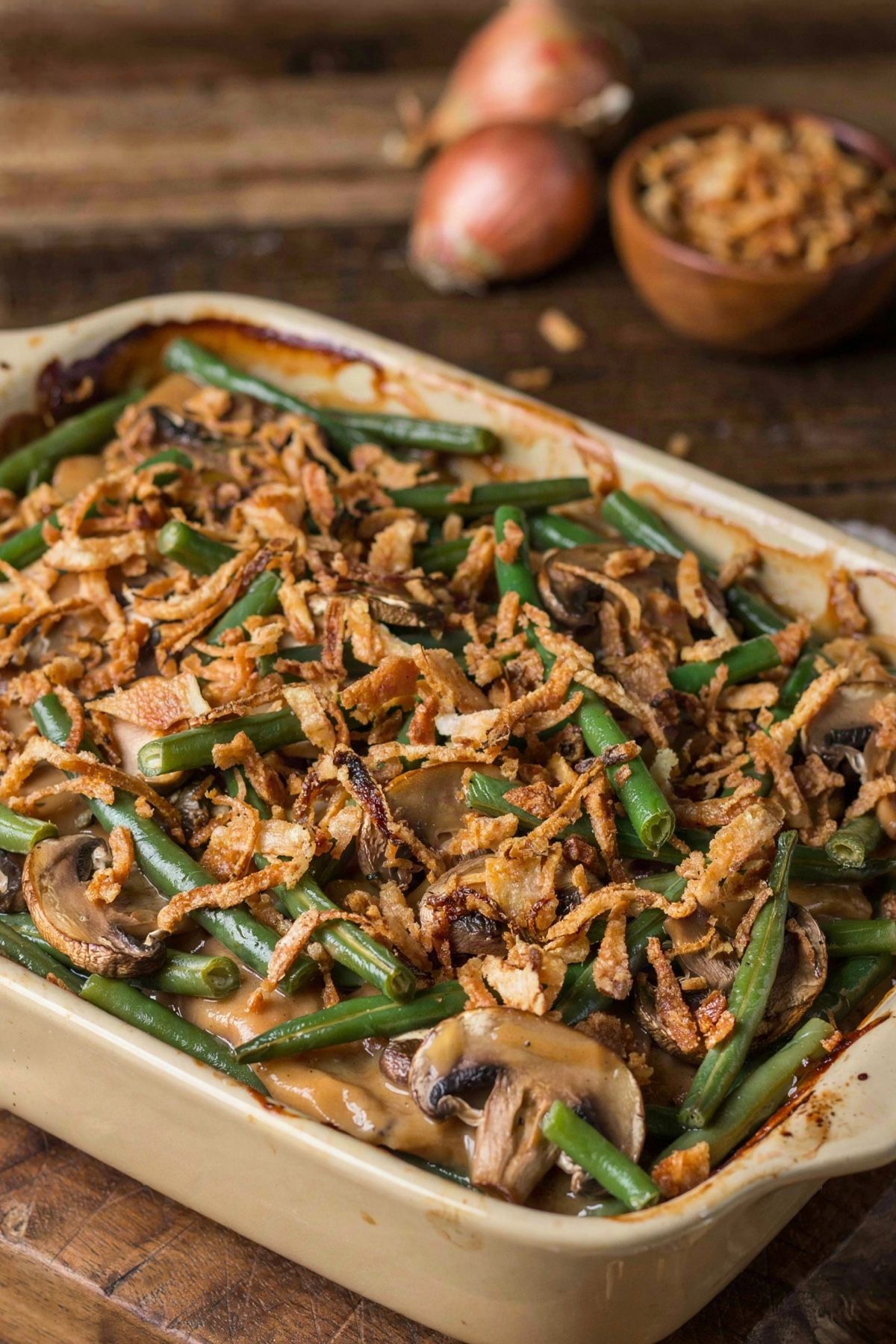 Vegan green bean casserole in a cream stoneware baking dish, cut green beans and sliced browned mushrooms in a thick tan mushroom sauce under a heaped layer of deep golden crispy fried onions.