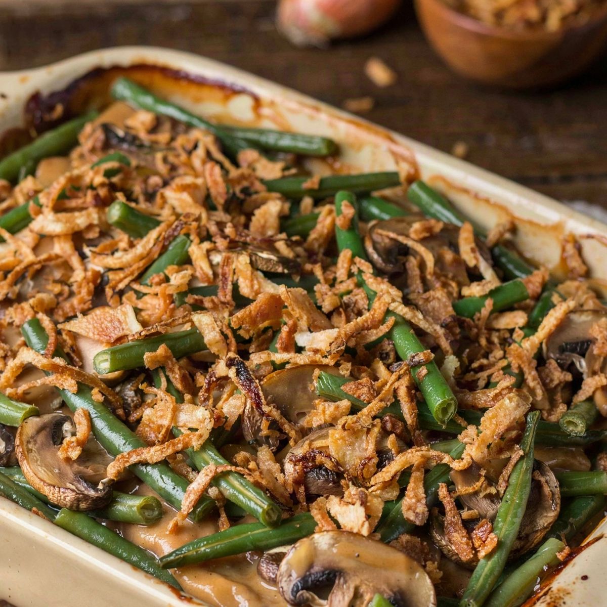 Vegan green bean casserole in a cream stoneware baking dish, cut green beans and sliced browned mushrooms in a thick tan mushroom sauce under a heaped layer of deep golden crispy fried onions.