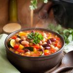A bowl of bean and vegetable soup with carrots, zucchini, bell peppers, and beans, garnished with parsley, on a rustic kitchen table.