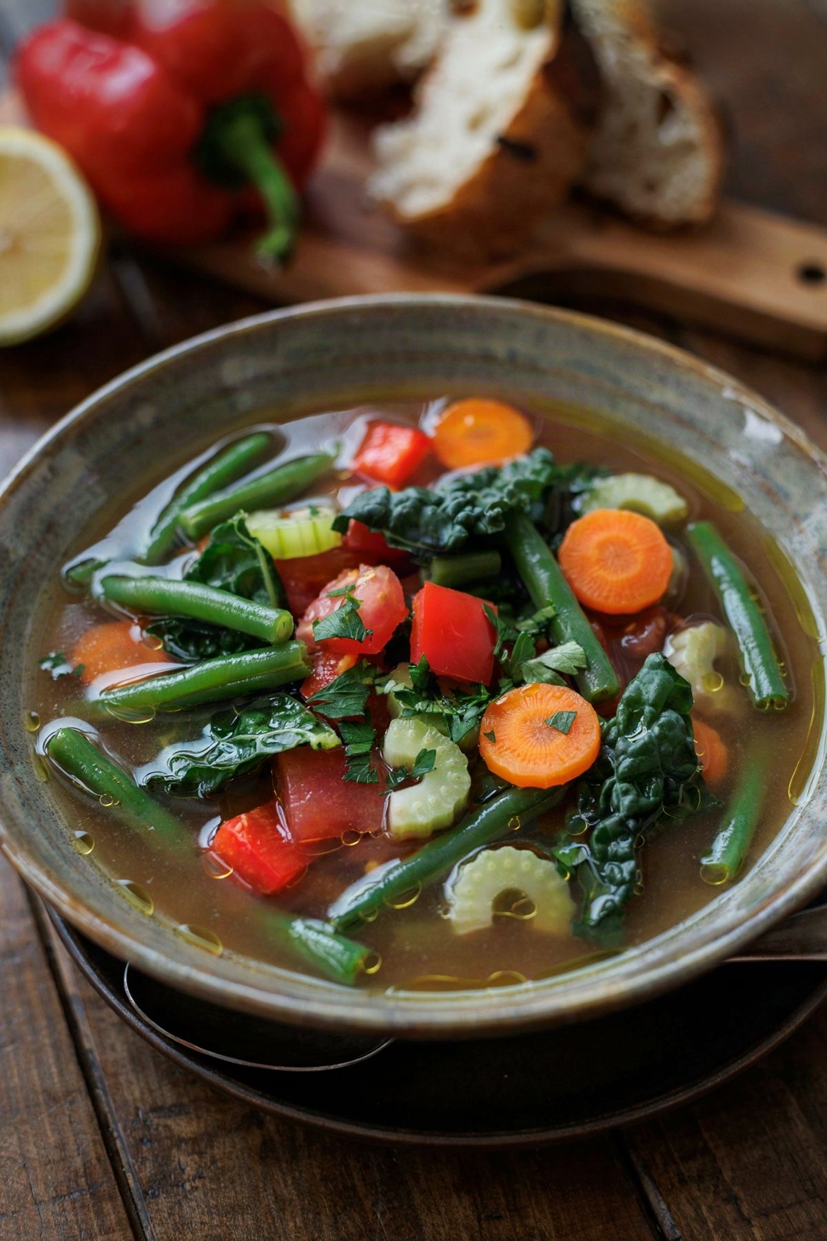 A bowl of brothy vegetable soup with green beans, carrots, red pepper and kale, with crusty bread alongside on a wooden table.