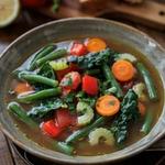 A bowl of brothy vegetable soup with green beans, carrots, red pepper and kale, with crusty bread alongside on a wooden table.