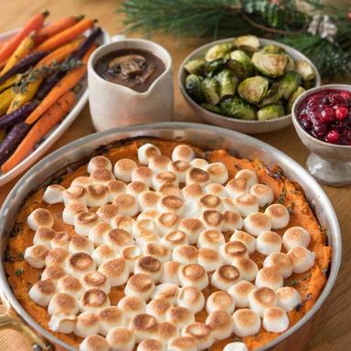 A vegetarian Thanksgiving table with sweet potato casserole under toasted marshmallows, a jug of mushroom gravy, roasted Brussels sprouts, cranberry sauce and roasted rainbow carrots.