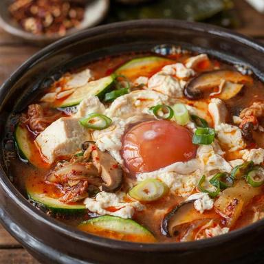 Vegetarian sundubu jjigae in a dark earthenware bowl, soft curds of silky tofu in a vivid red chili broth with zucchini, kimchi and shiitake, one soft-yolked egg in the center and sliced scallions on top.